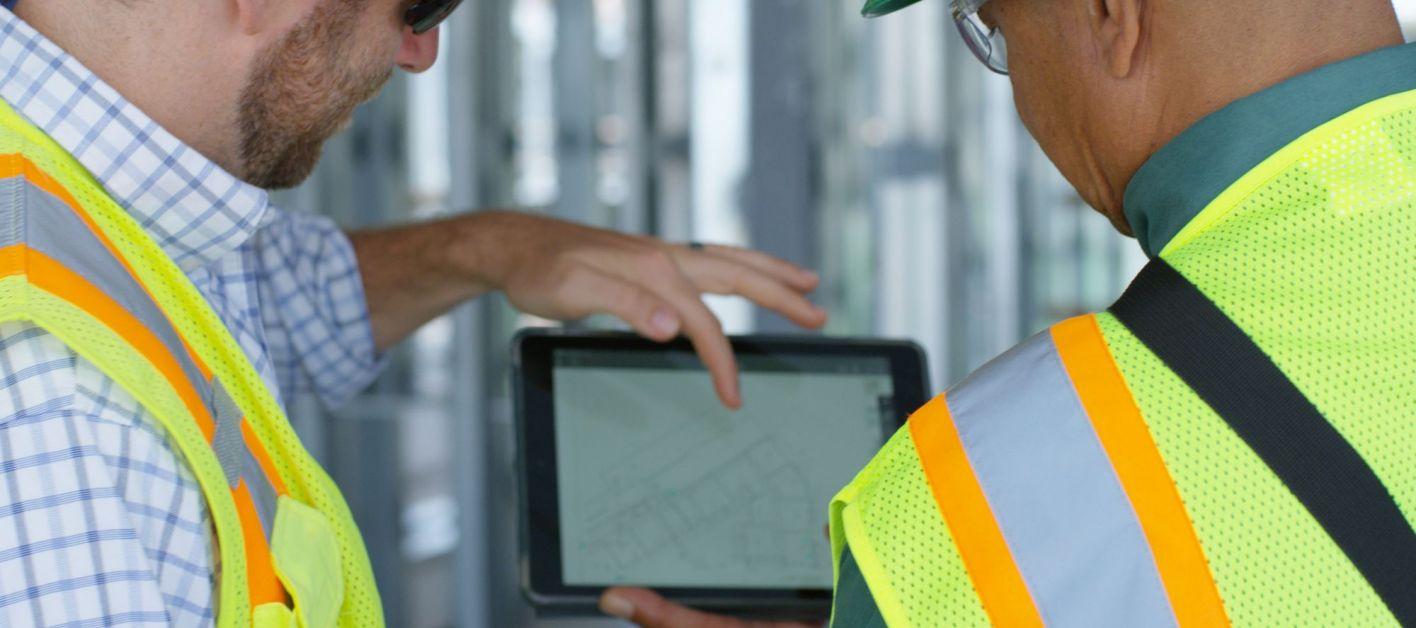 Two construction workers in safety vests inspect a tablet together.