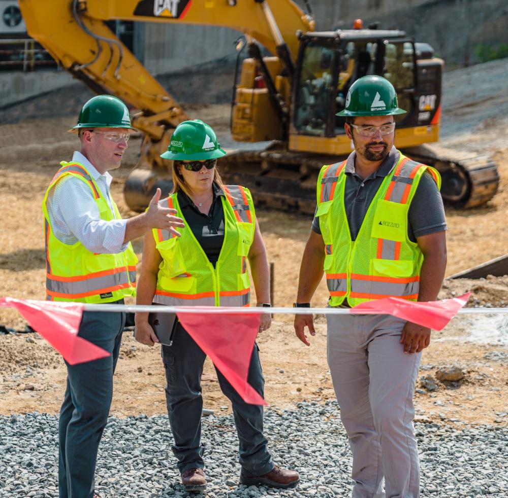 Three construction workers in safety vests and helmets discuss near machinery on a work site.