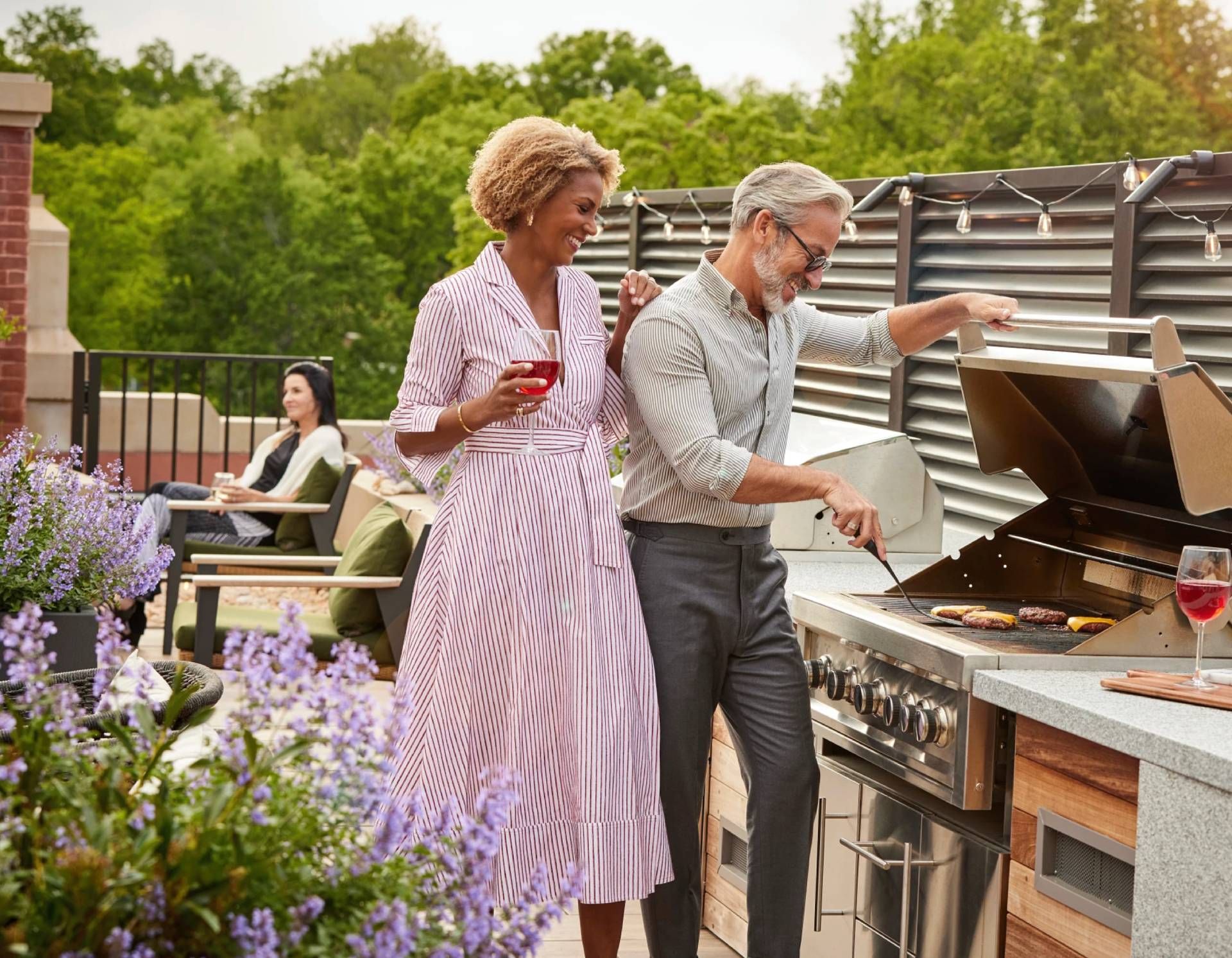 A couple shares a moment while grilling outdoors, with a guest relaxing nearby.
