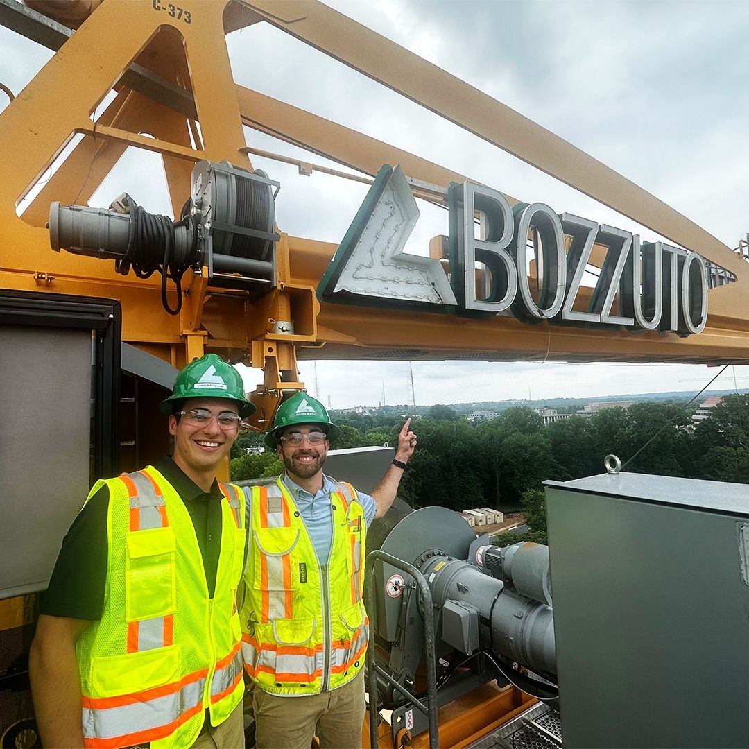 Two workers in safety vests and helmets stand next to a crane with a "BOZZUTO" sign.