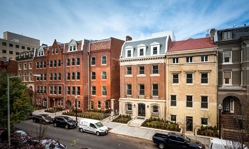 Row of brick and stone buildings along a tree-lined street under a blue sky.