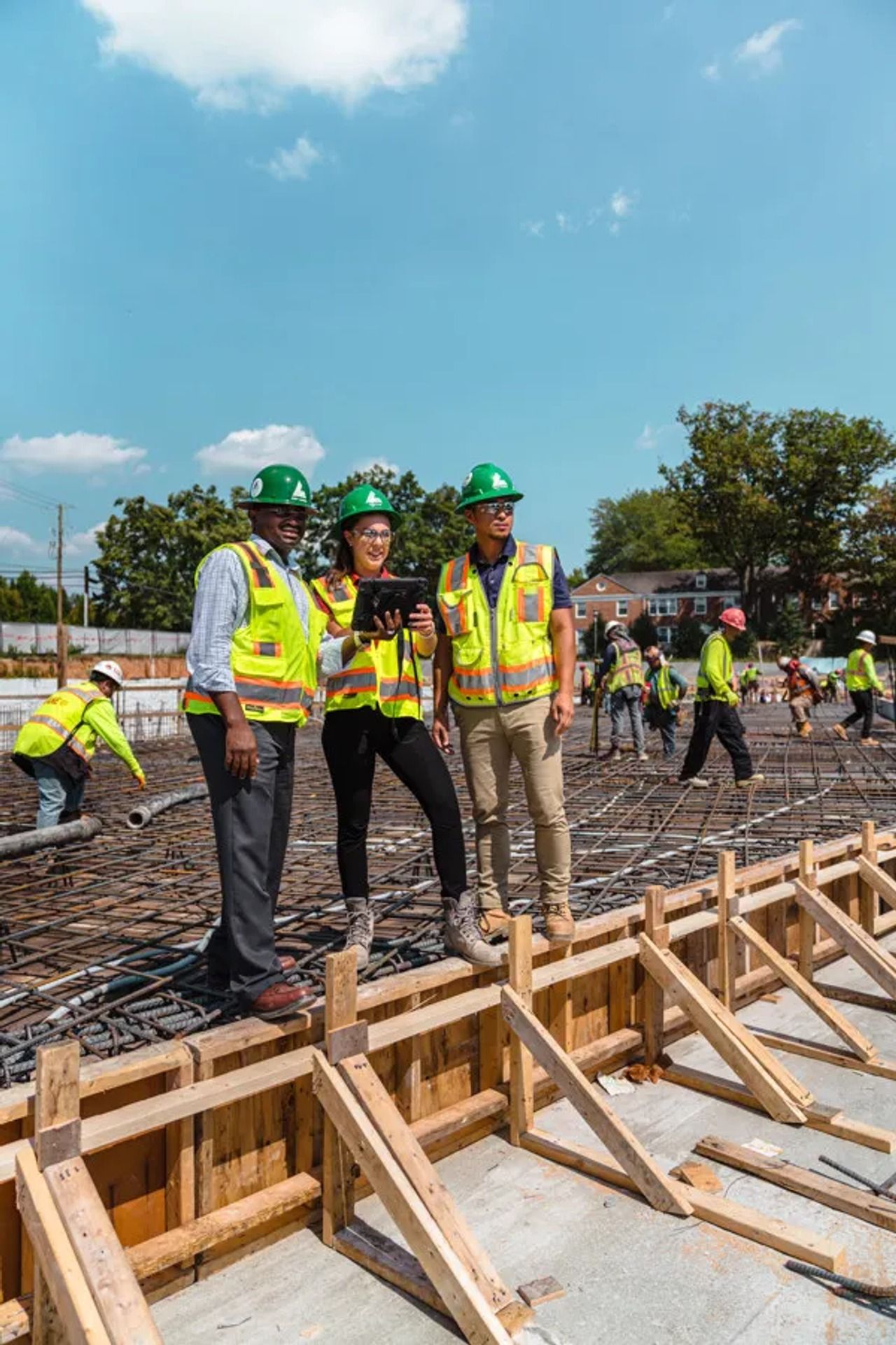 Three construction workers in hard hats and vests on a building site, with others working behind.