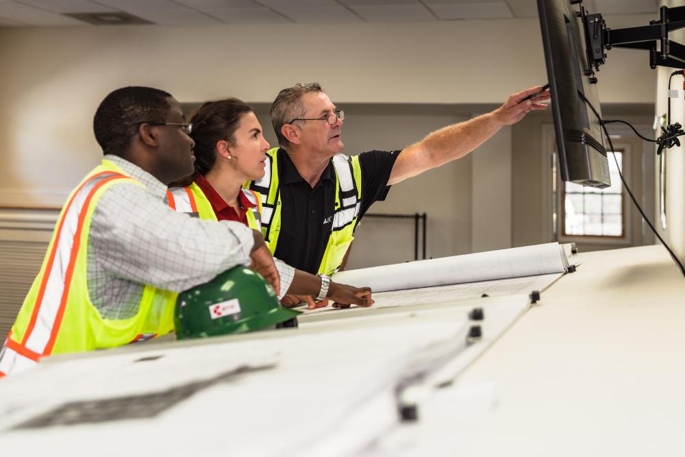 Three construction professionals review plans at a table, pointing at a screen.