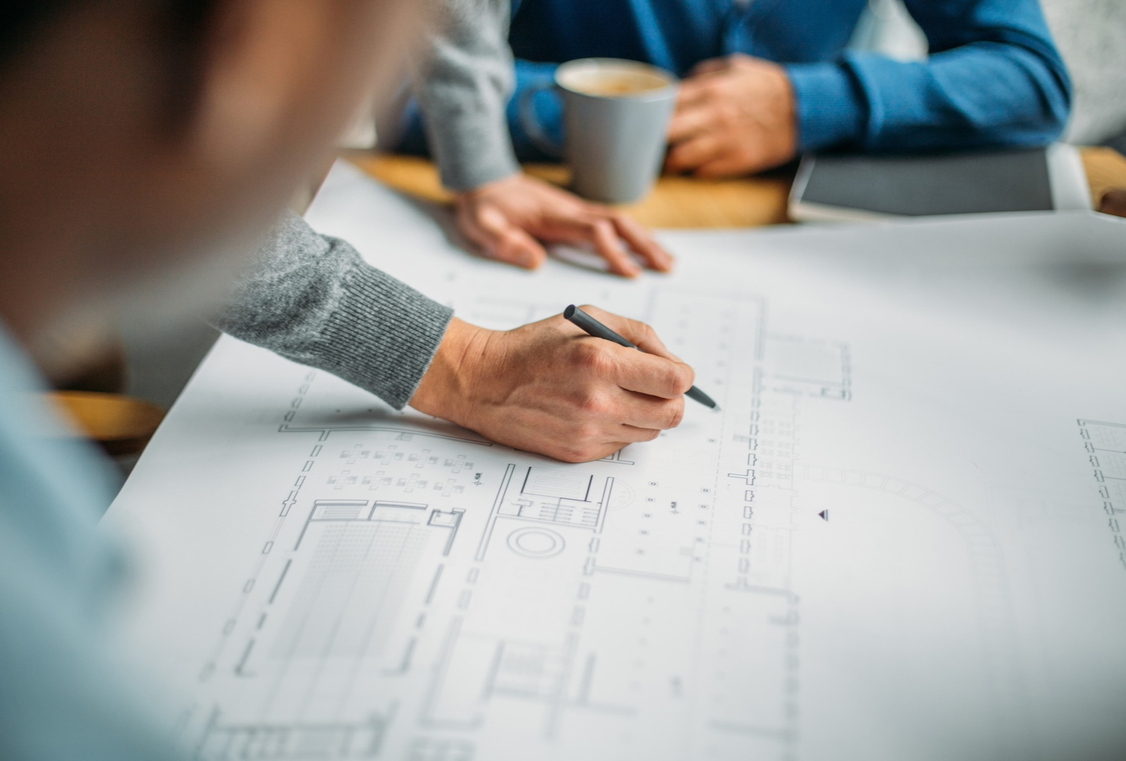 Person drawing on architectural plans with others and a coffee cup in the background.