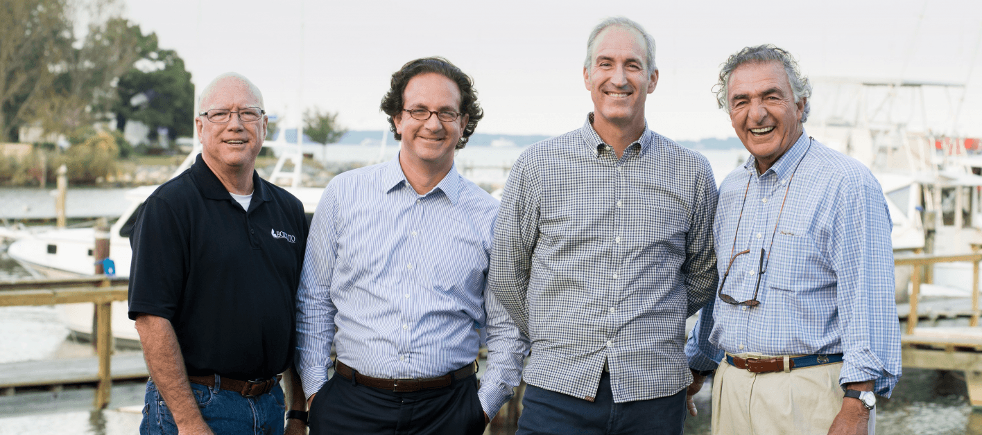 Four men standing together by water, smiling on a wooden dock.