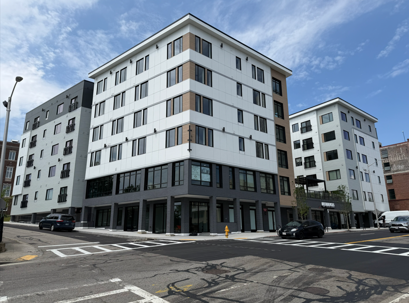 Modern mid-rise apartment buildings with white, gray, and wood-accented exteriors at a city street intersection.