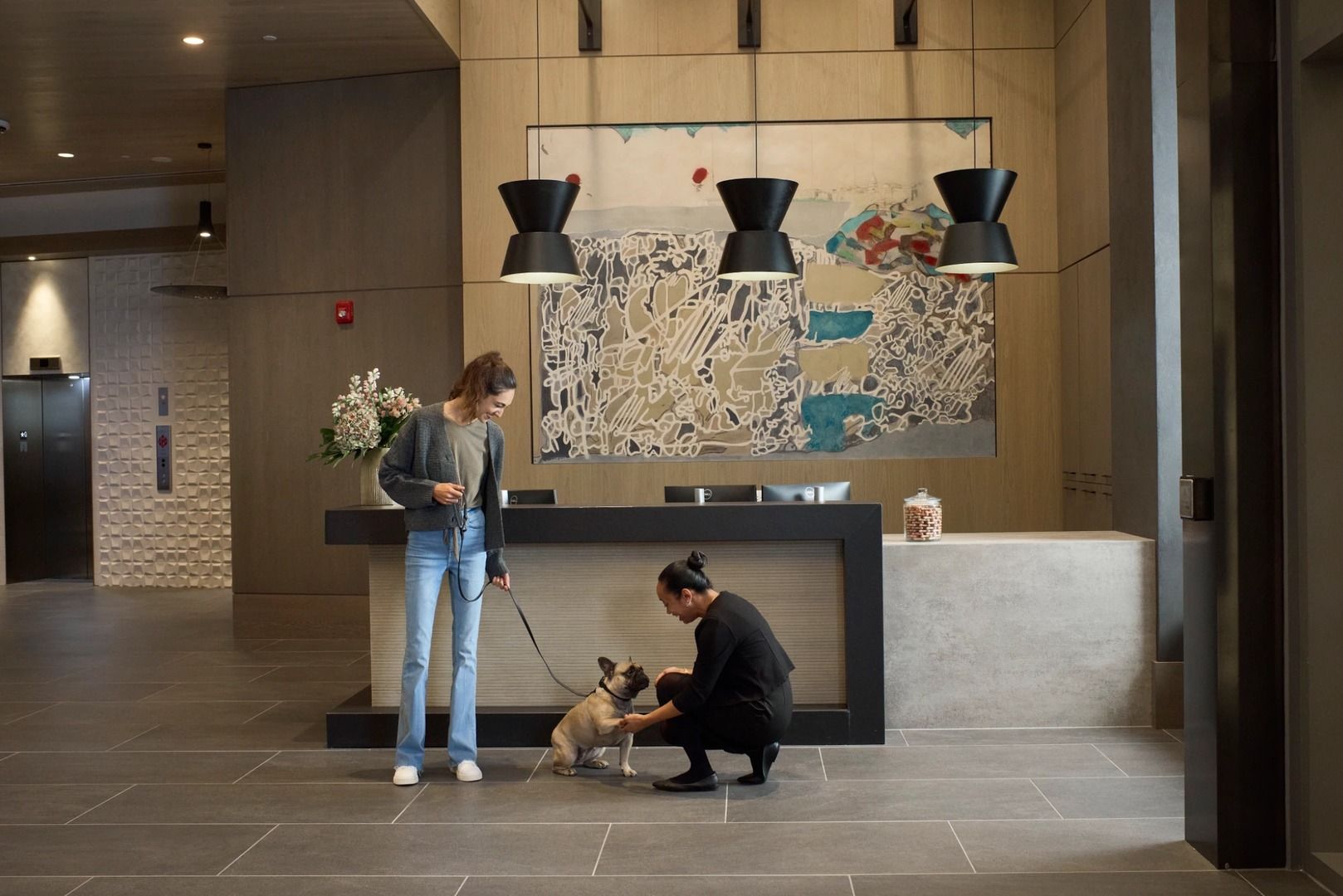 A woman stands with a dog while a man crouches to pet it in a modern lobby.
