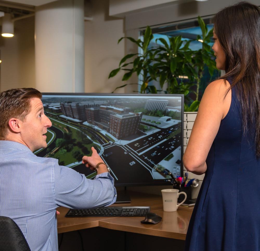 Two people discuss a building design while looking at a computer screen in an office setting.