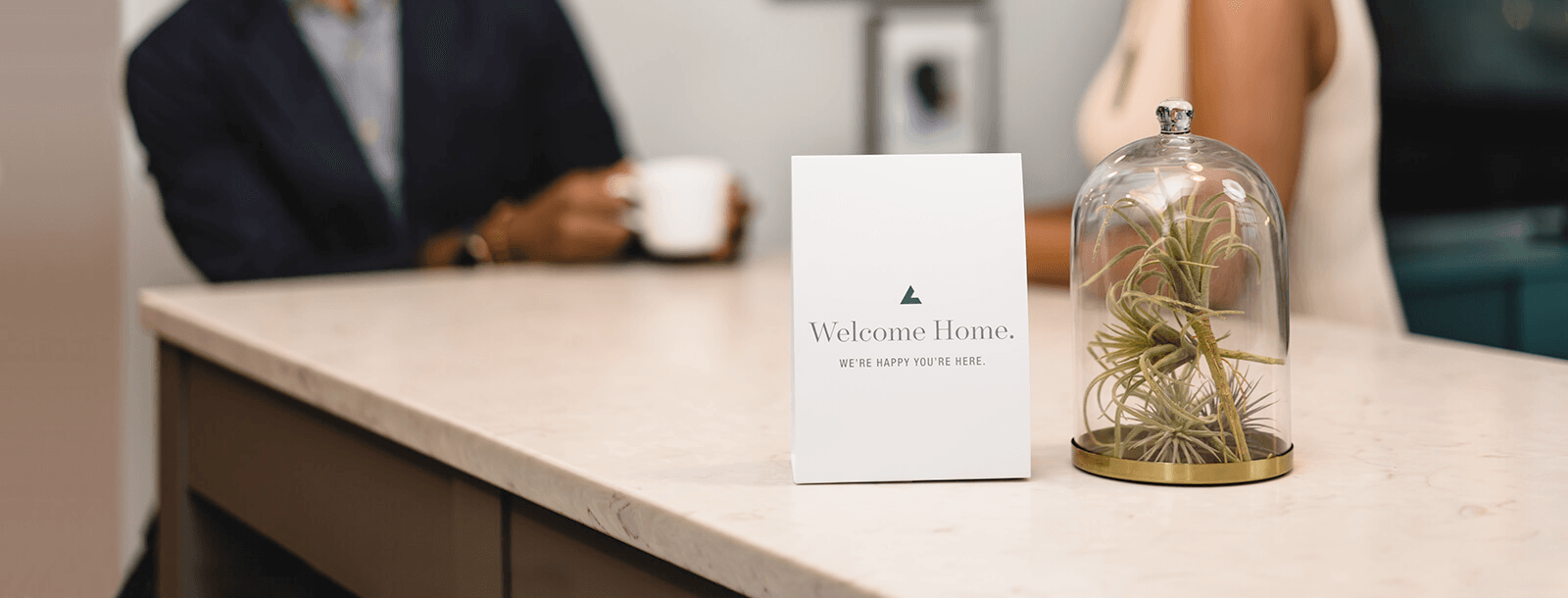 A welcome sign on a countertop with a glass cloche and a person holding a cup in the background.