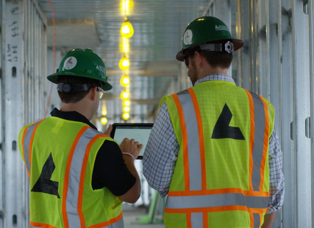 Two workers in safety vests and helmets examine a tablet in a hallway.