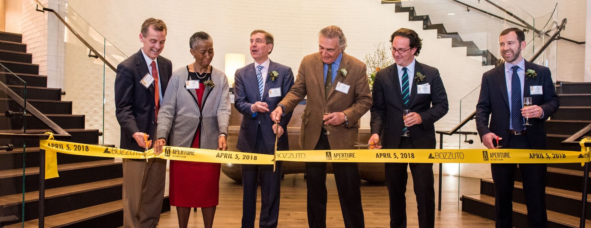 Group of six people cutting a yellow ribbon at a formal event indoors.