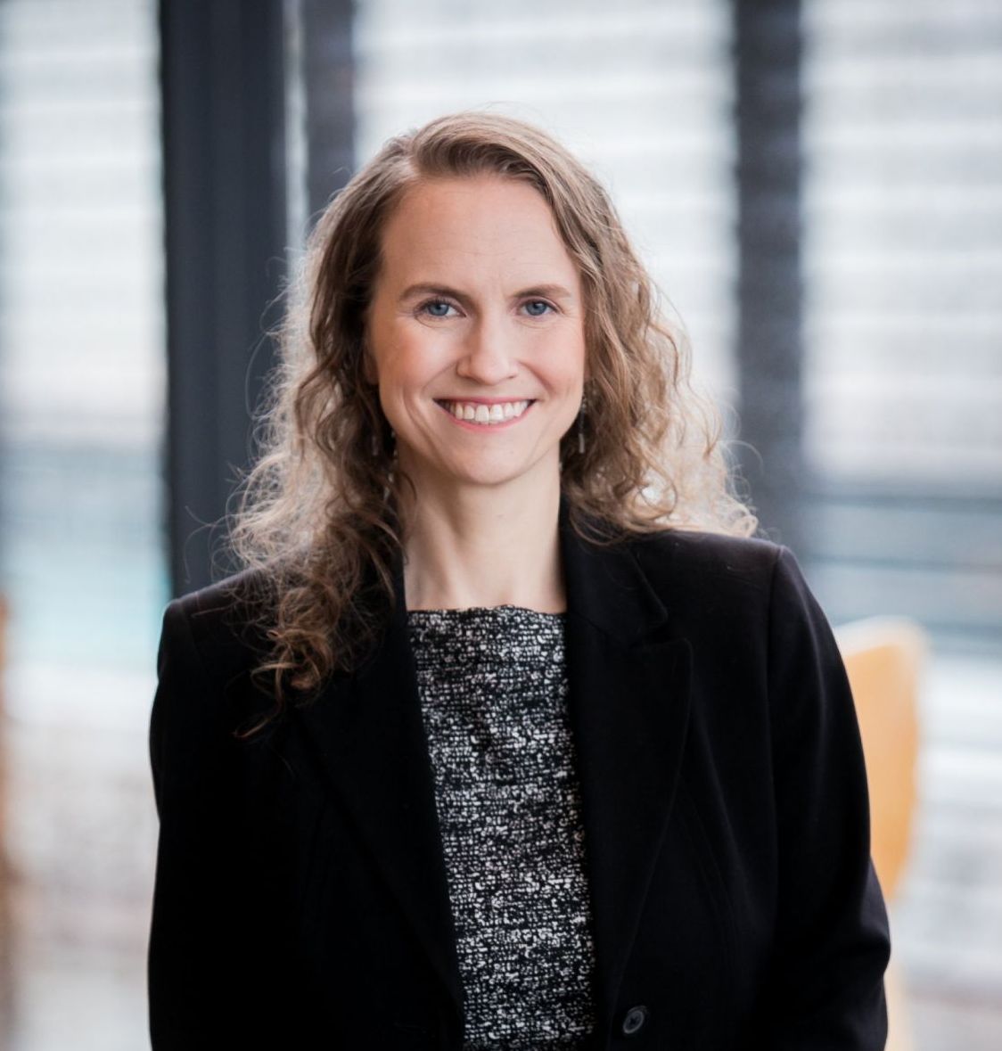 Smiling woman in a blazer stands in front of windowed background.