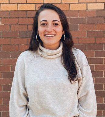 Smiling person in a gray sweater standing against a brick wall.