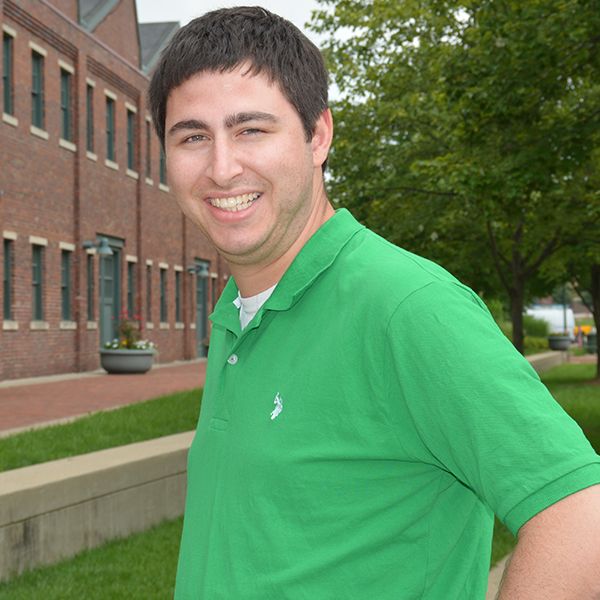 Smiling man in a green shirt standing outside by a brick building.