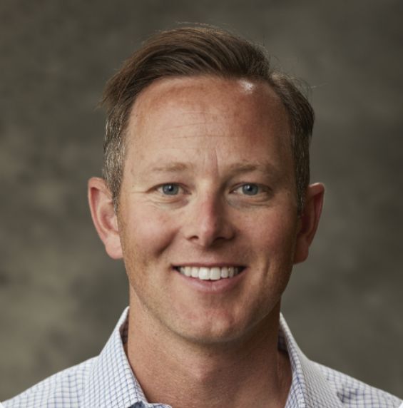 Smiling man with short hair, wearing a checkered shirt, against a textured background.