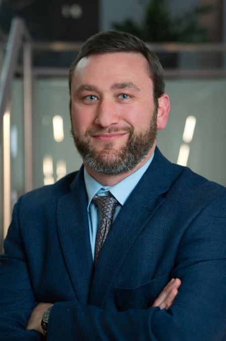 A man in a suit smiles, arms crossed, with a modern interior background.
