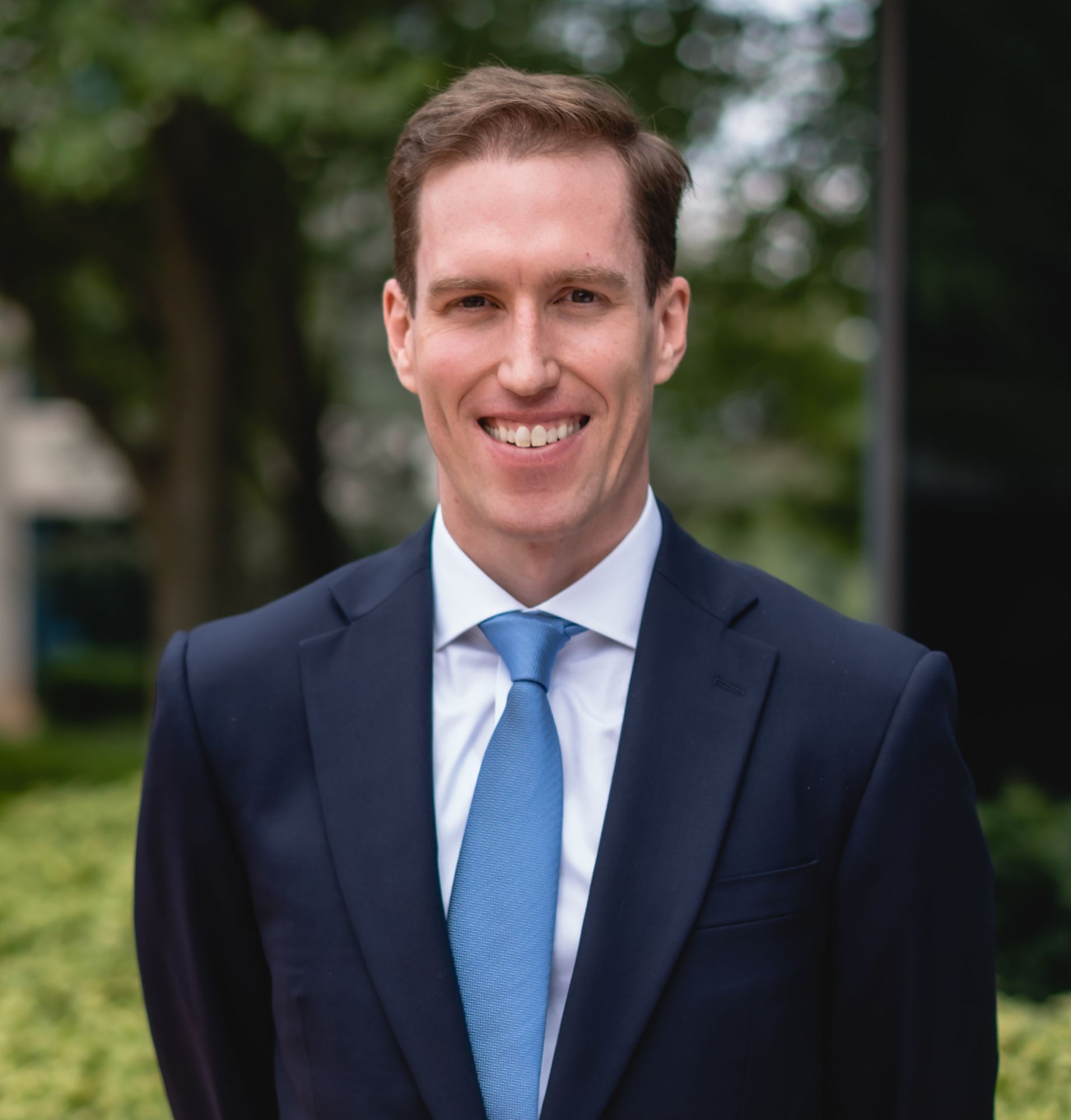 A man in a suit with a blue tie stands outdoors, smiling in a green environment.