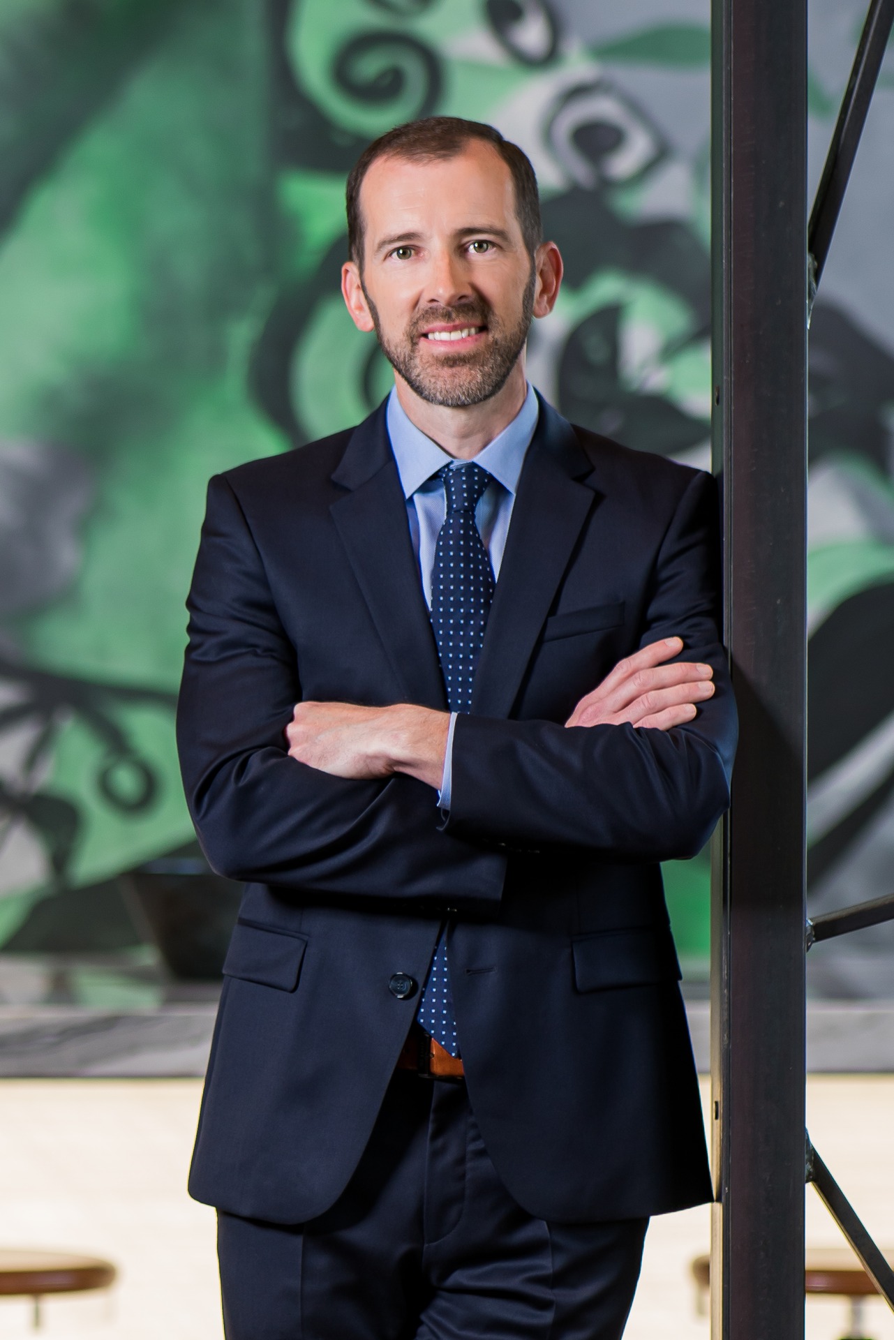 Portrait of Mike Henehan in a suit, standing with arms crossed against a colorful background.