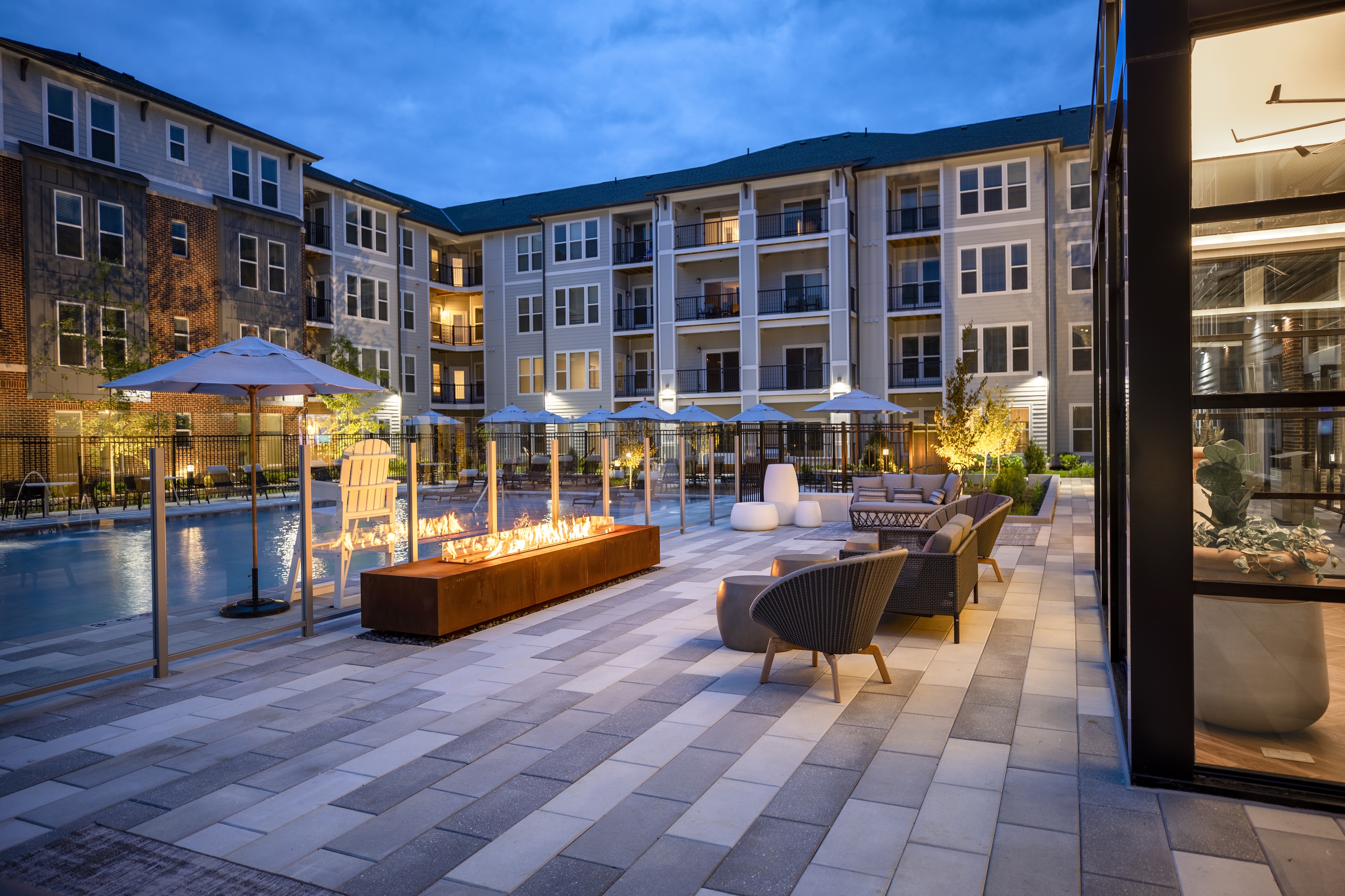 Poolside view of an apartment complex at dusk with lounge chairs and fire pits.