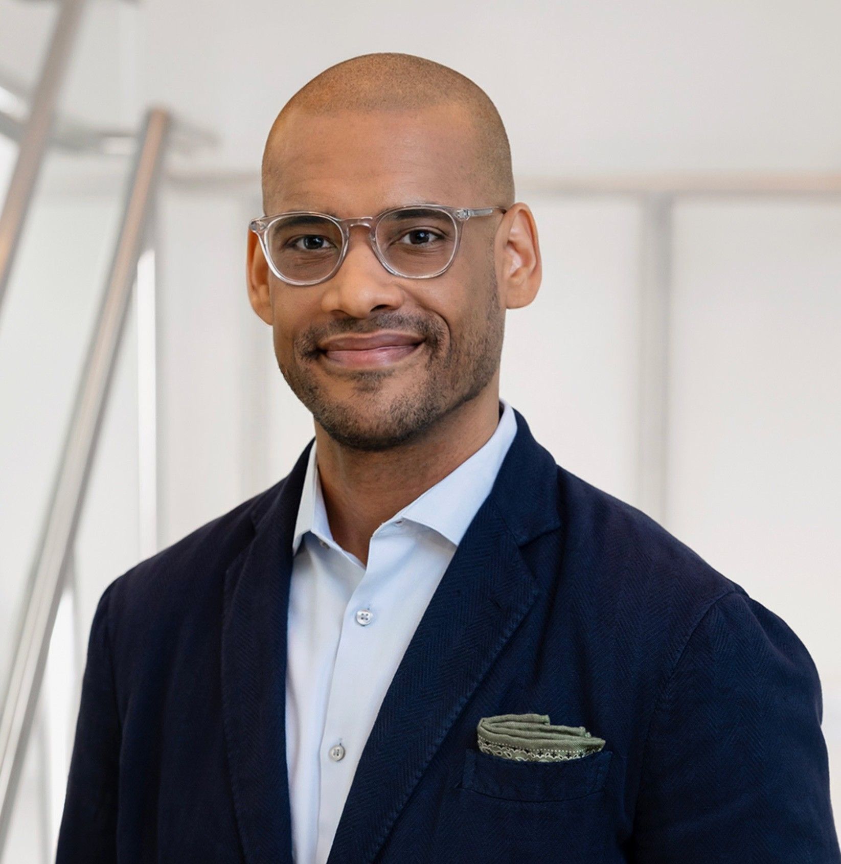 A smiling man in glasses wearing a dark blazer and light shirt, indoors with natural light.