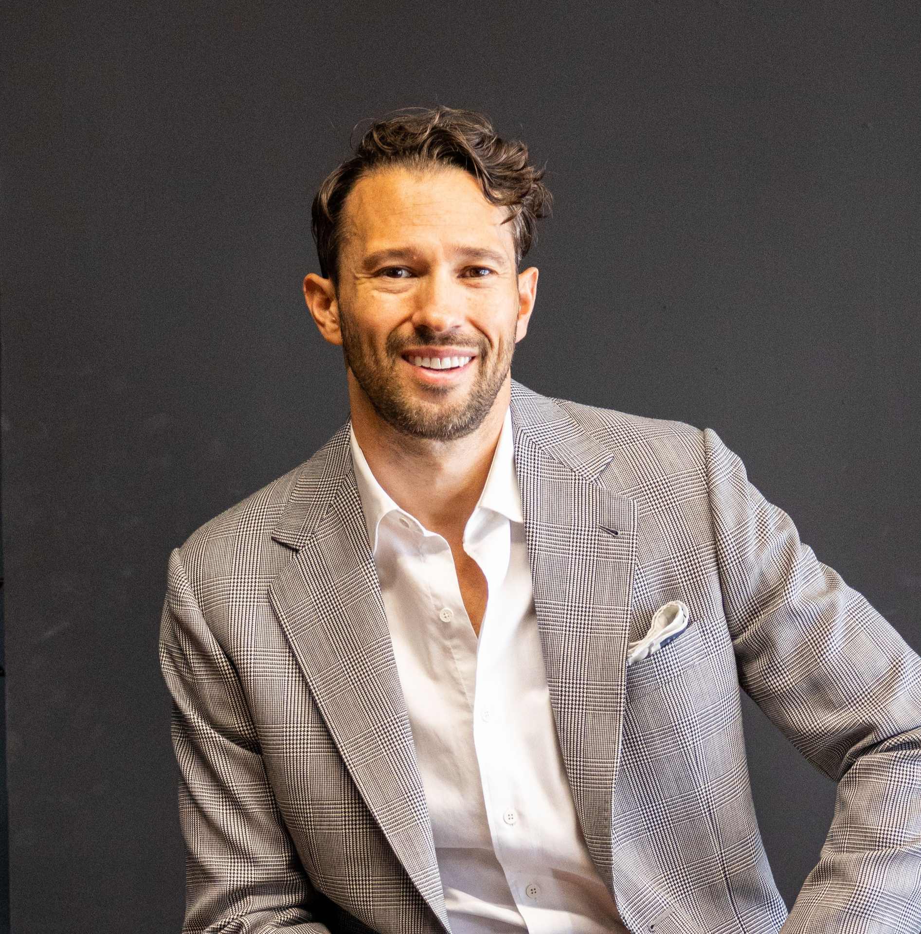 Nathan Veliz is smiling while wearing a light gray suit against a dark background.
