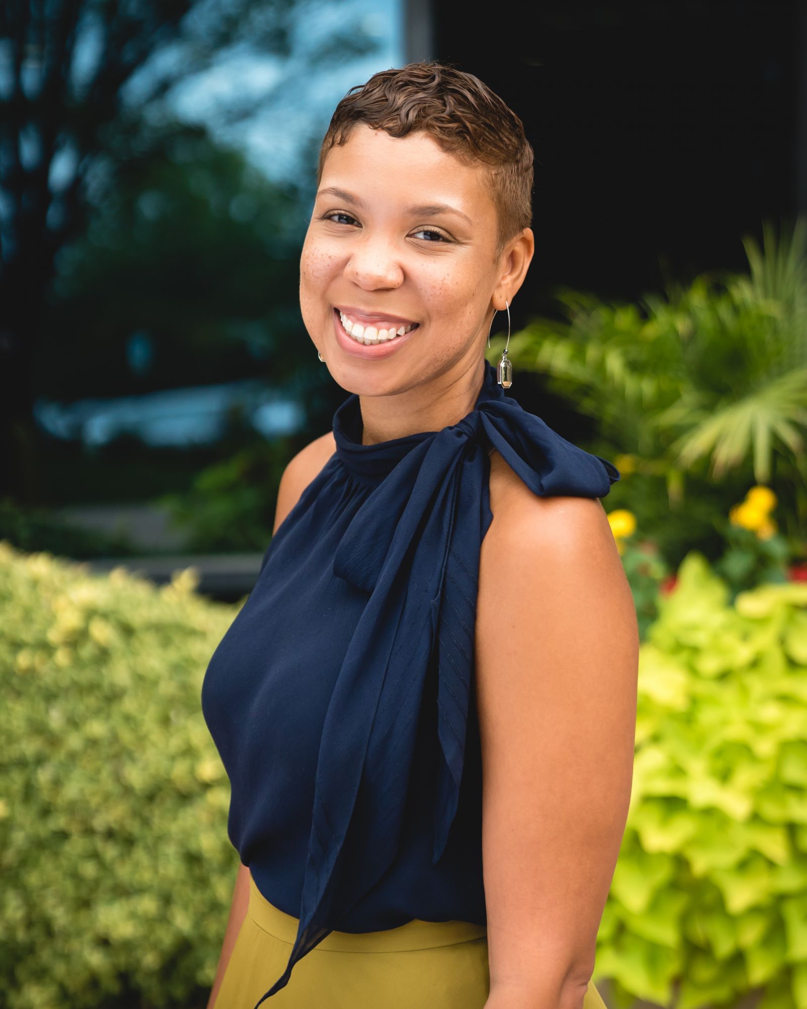 Smiling woman in a navy top stands near green plants.