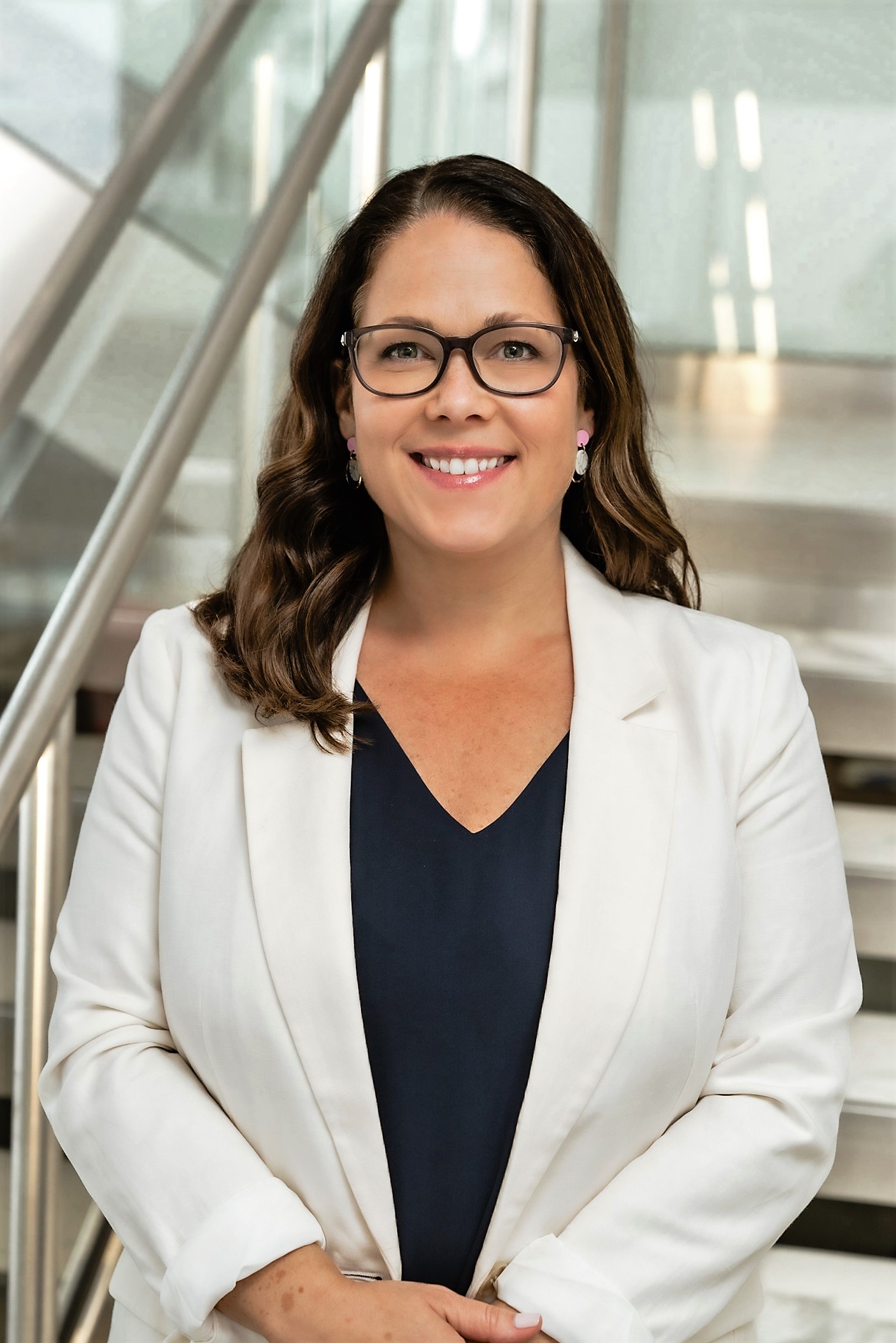 Professional portrait of Amy Medugno, smiling, wearing a white blazer, in a modern interior setting.