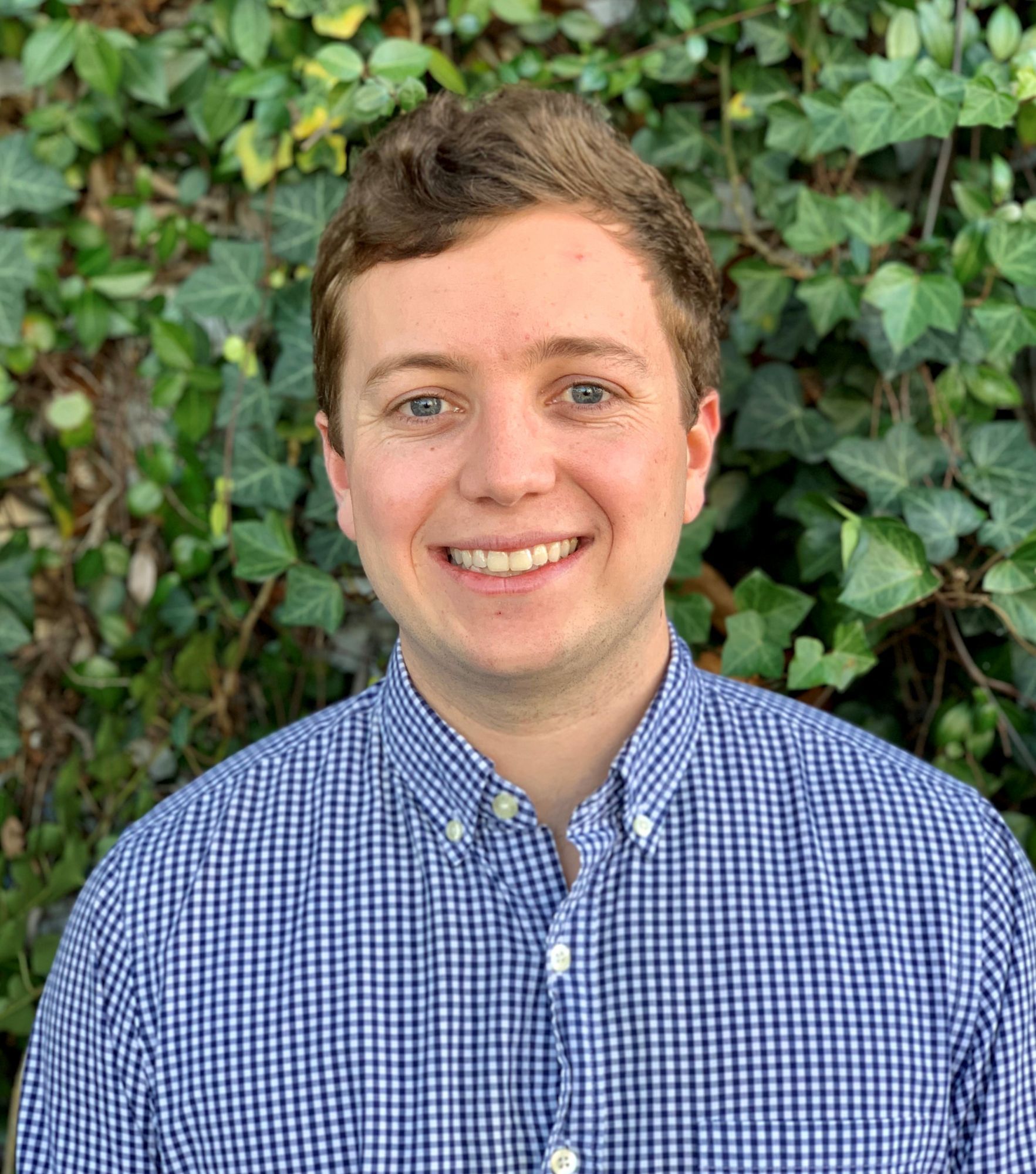 Smiling person in a blue checkered shirt against a green leafy background.