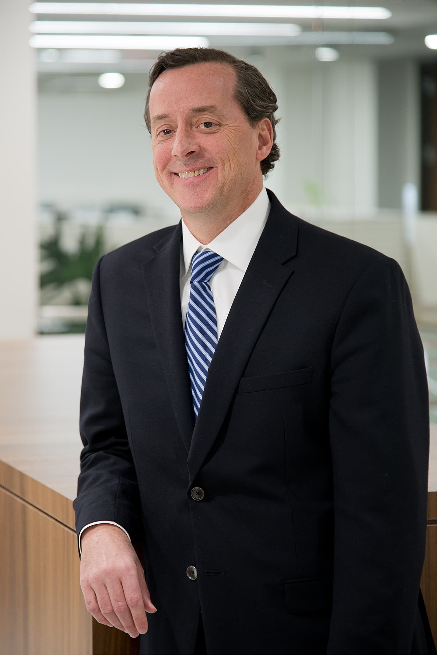 A man in a suit stands beside a light-colored wall, smiling in an office setting.