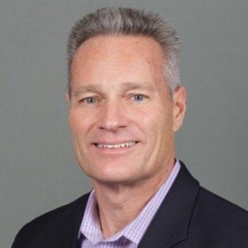 A professional man with gray hair smiling, wearing a suit against a gray background.
