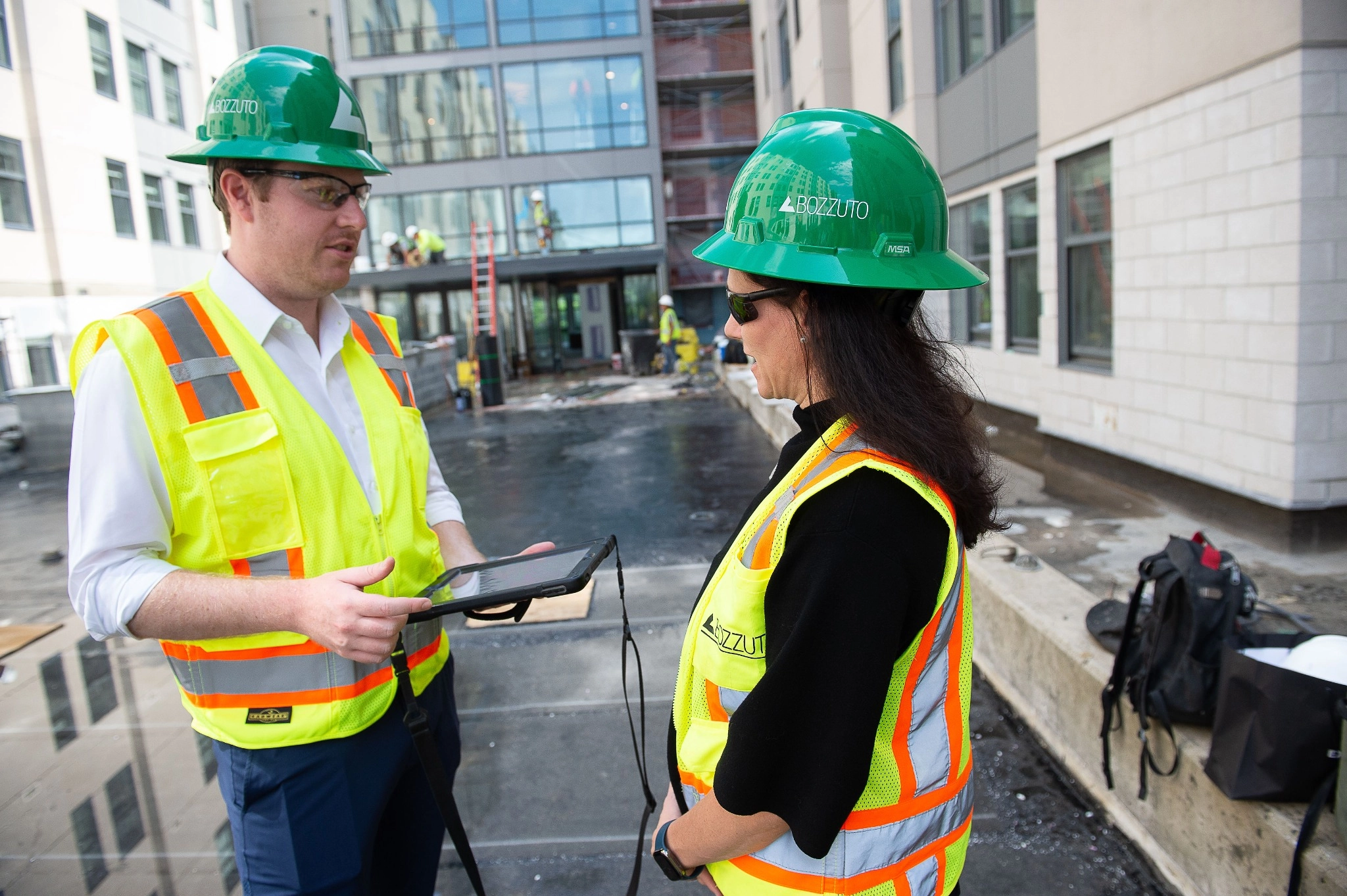 Two construction workers in green hard hats discuss plans on a tablet.