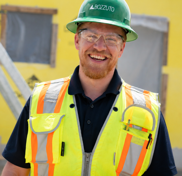 Smiling construction worker wearing safety glasses, a green Bozzuto hard hat, and a high-visibility vest at a jobsite.