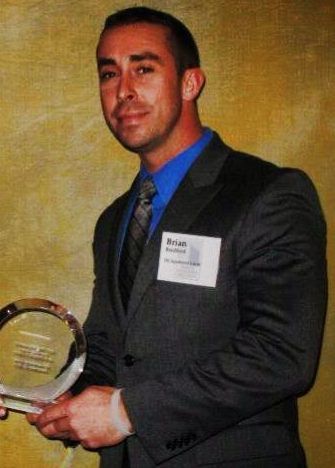 A man in a suit holding an award, standing against a textured background.