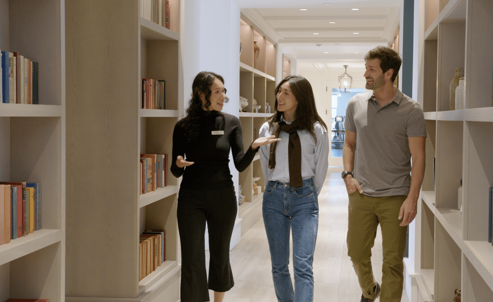 Three people walk and chat in a well-lit library aisle filled with books.