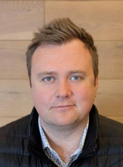 A man with short hair wearing a black vest, standing in front of a wooden background.