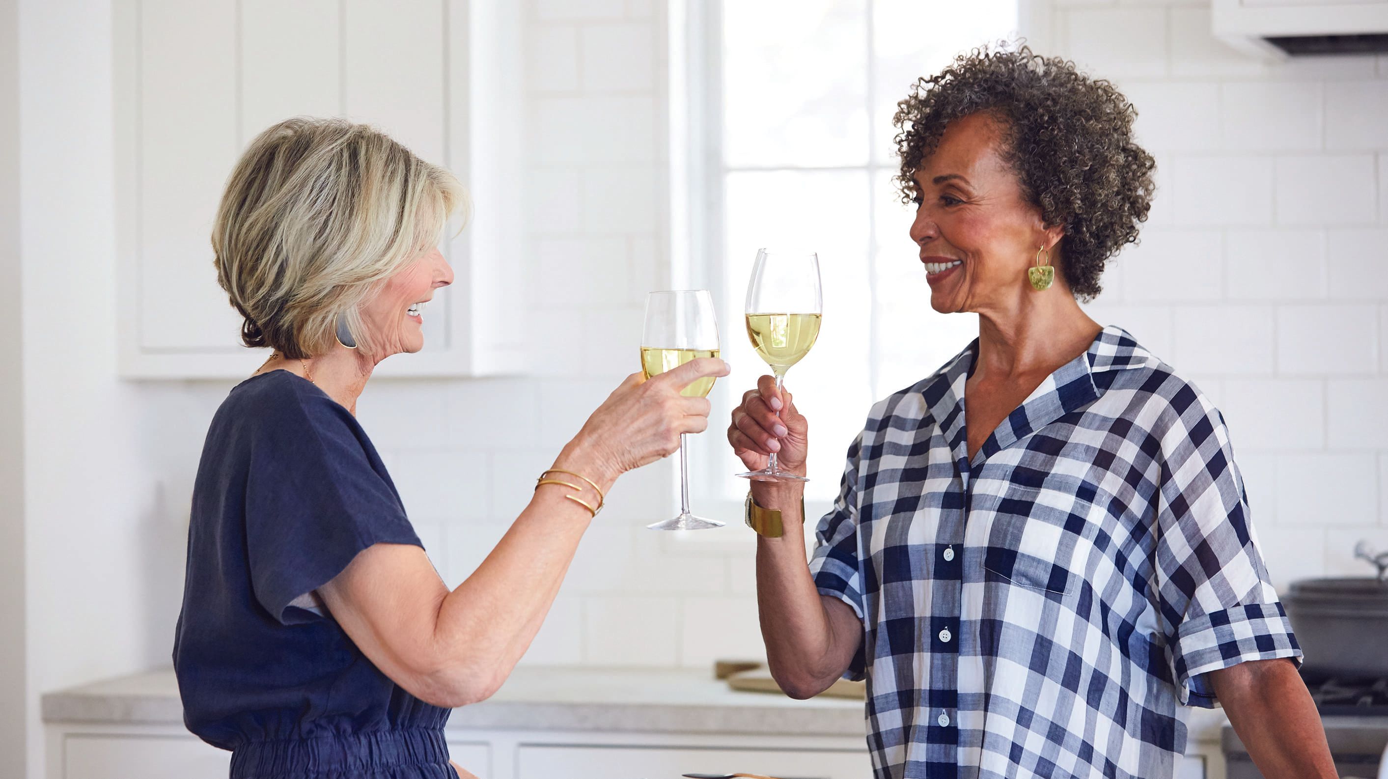Two women toasting with glasses of wine in a bright kitchen.