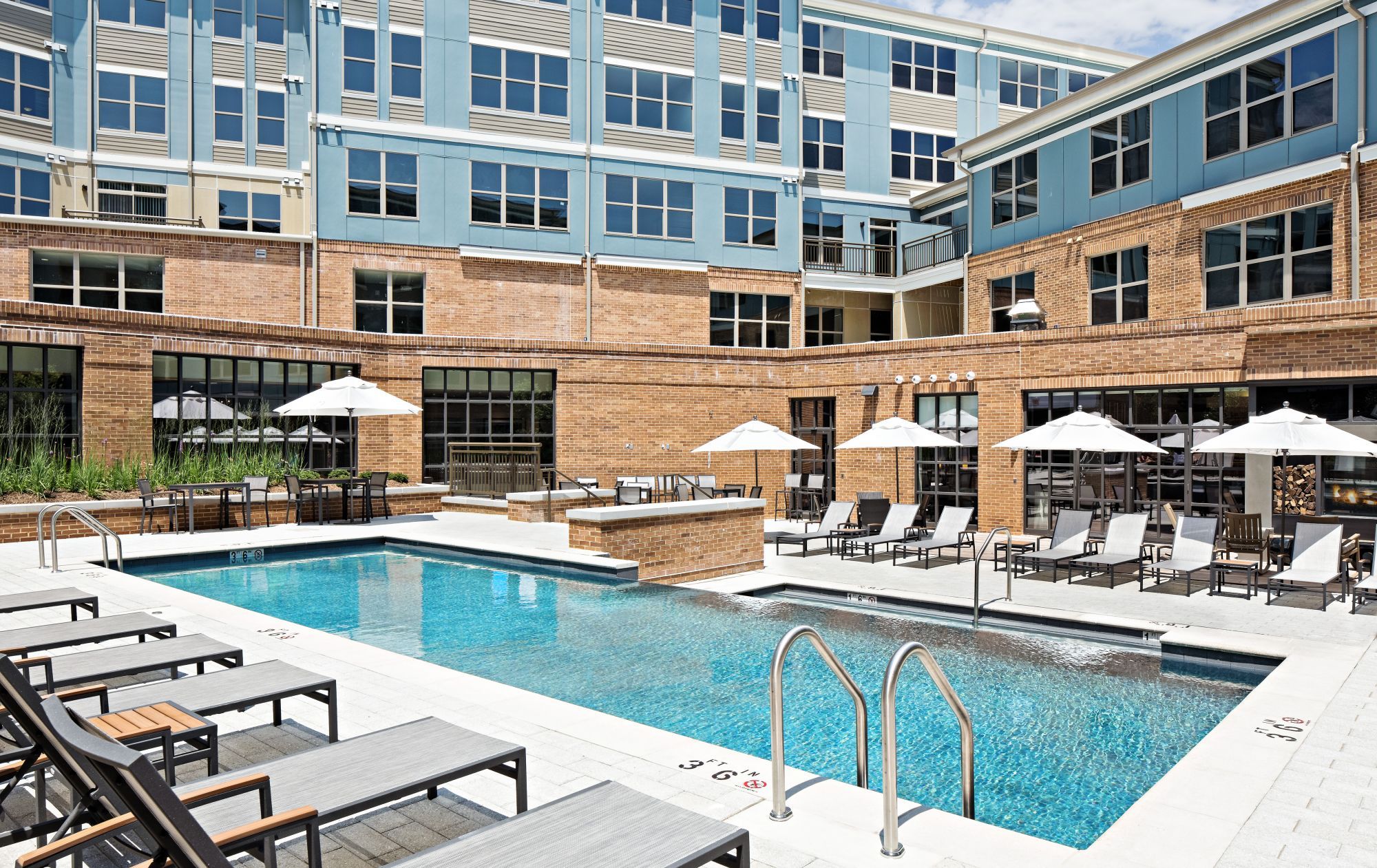 Modern apartment complex with a pool, deck chairs, and umbrellas in a sunny courtyard.
