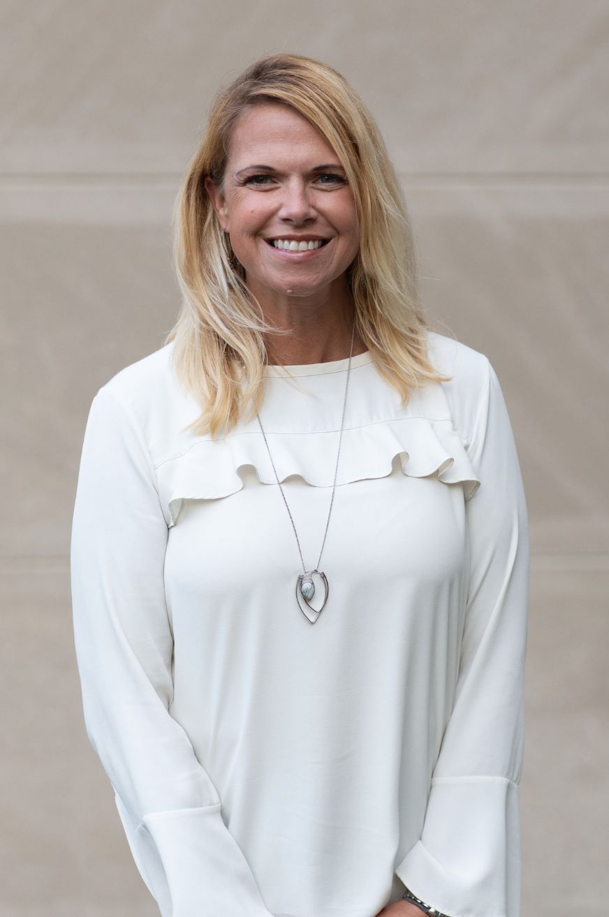 Smiling woman with long blonde hair, wearing a white top and standing against a neutral background.