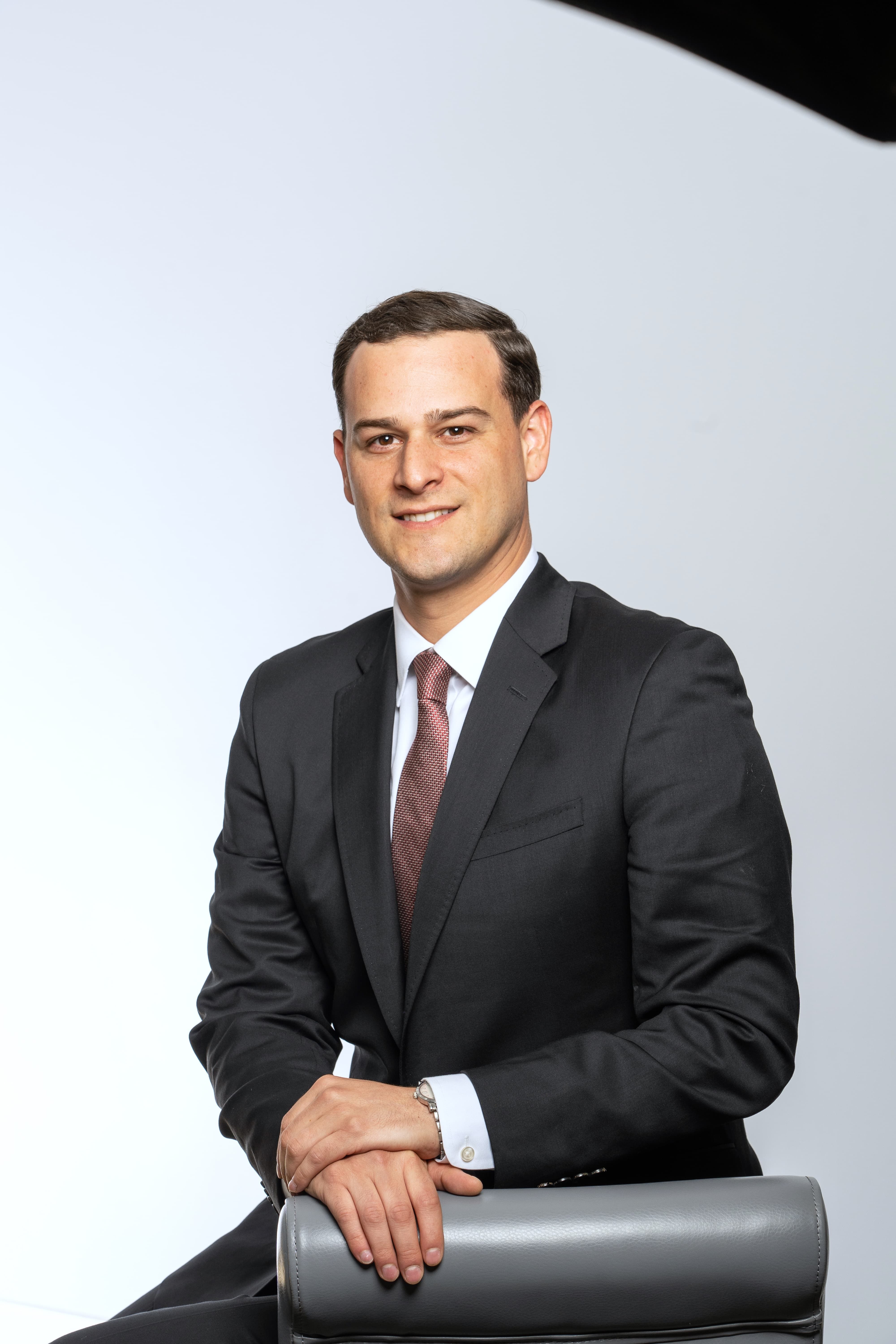 A professionally dressed man sitting on a chair with a neutral background.