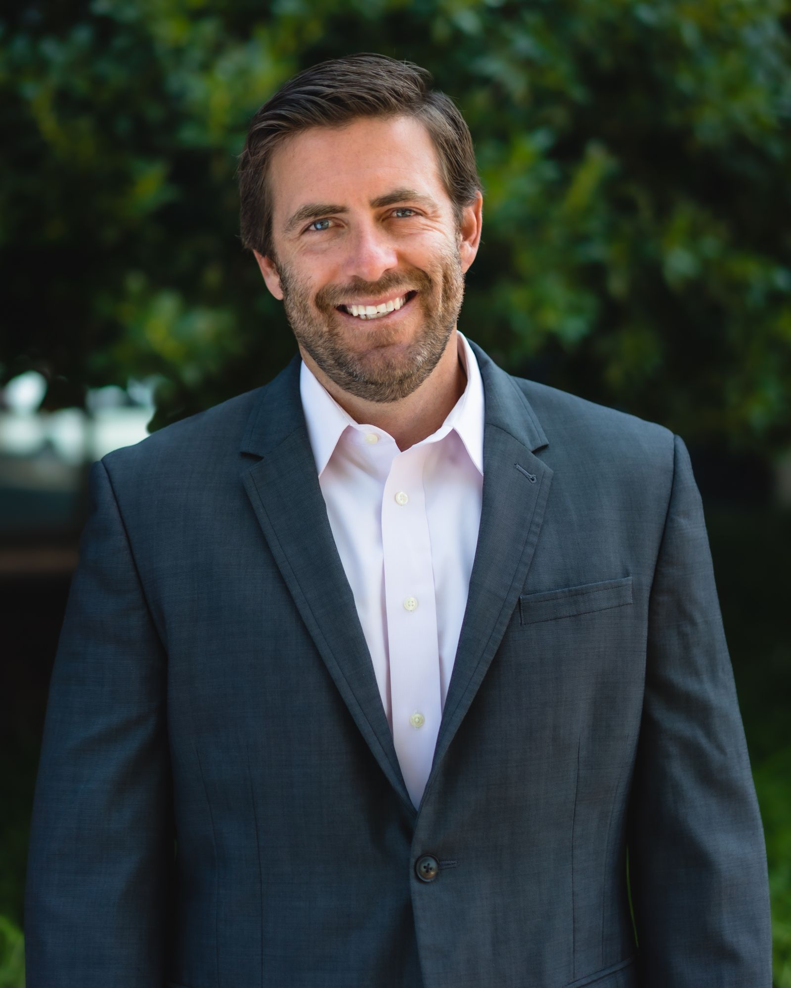 Smiling man in a suit standing outdoors with greenery in the background.