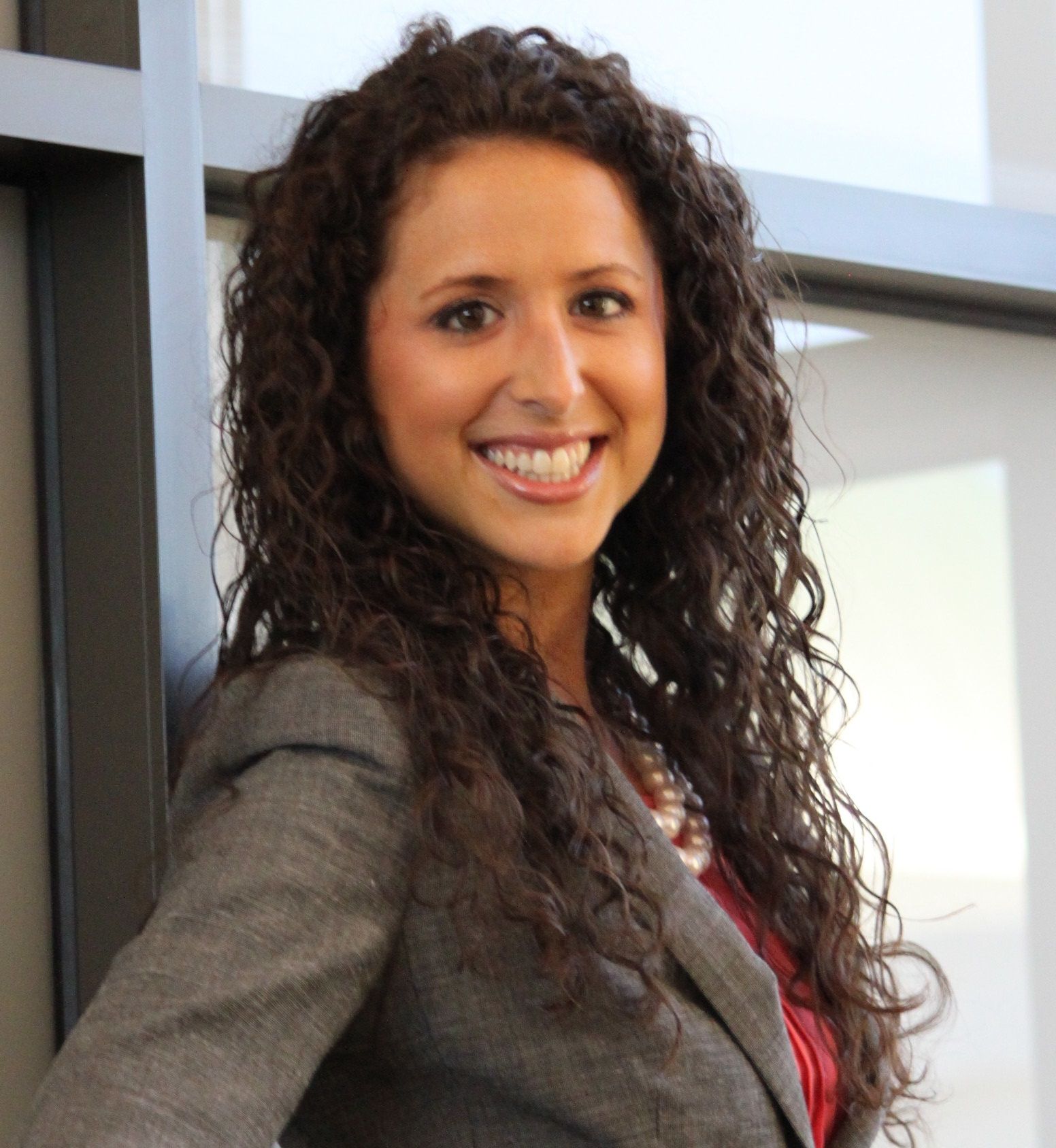 Smiling woman with curly hair in a professional outfit standing by a window.