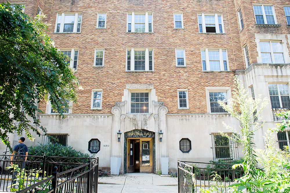 Brick apartment building with a central entrance and multiple windows, surrounded by greenery.