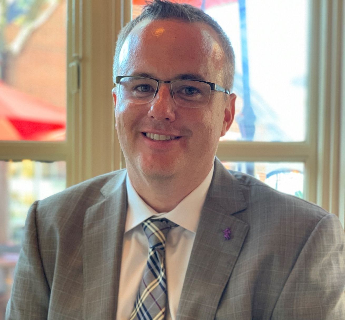 A smiling man in a suit and glasses is seated by a window.