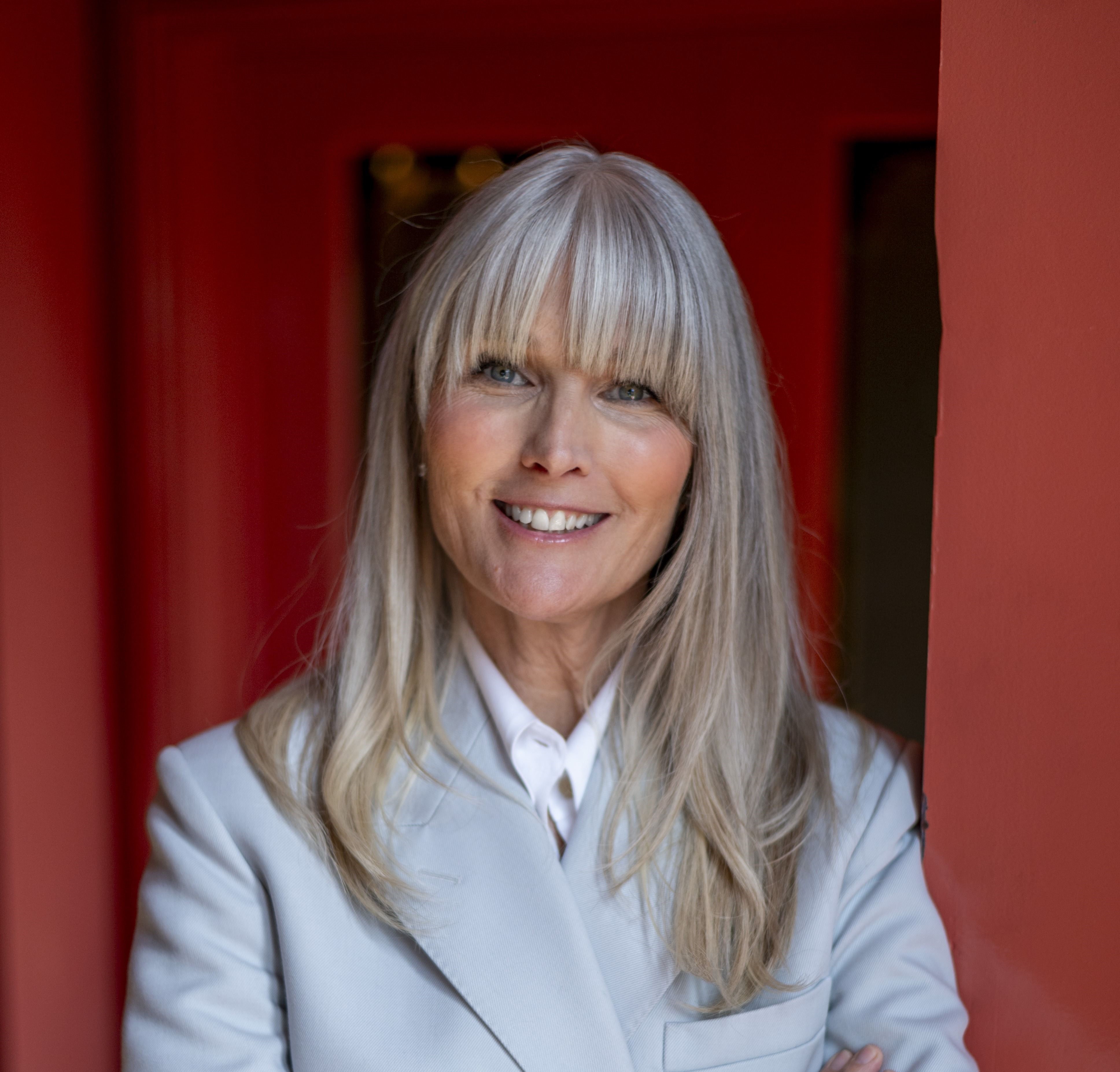 Heather Wallace smiles while standing in a brightly colored doorway, wearing a light-colored suit.