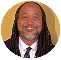 A smiling man with long dreadlocks wearing a suit against a yellow background.