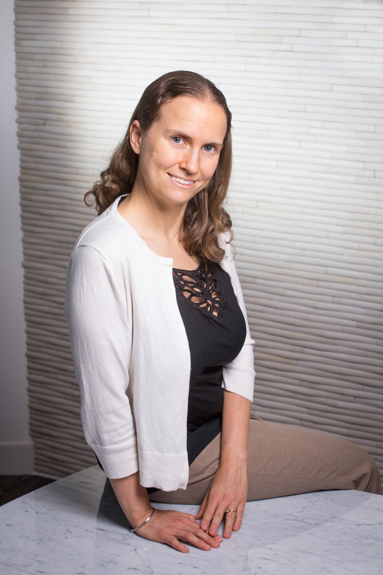 Smiling woman in a white cardigan poses with hands on a table in a well-lit space.