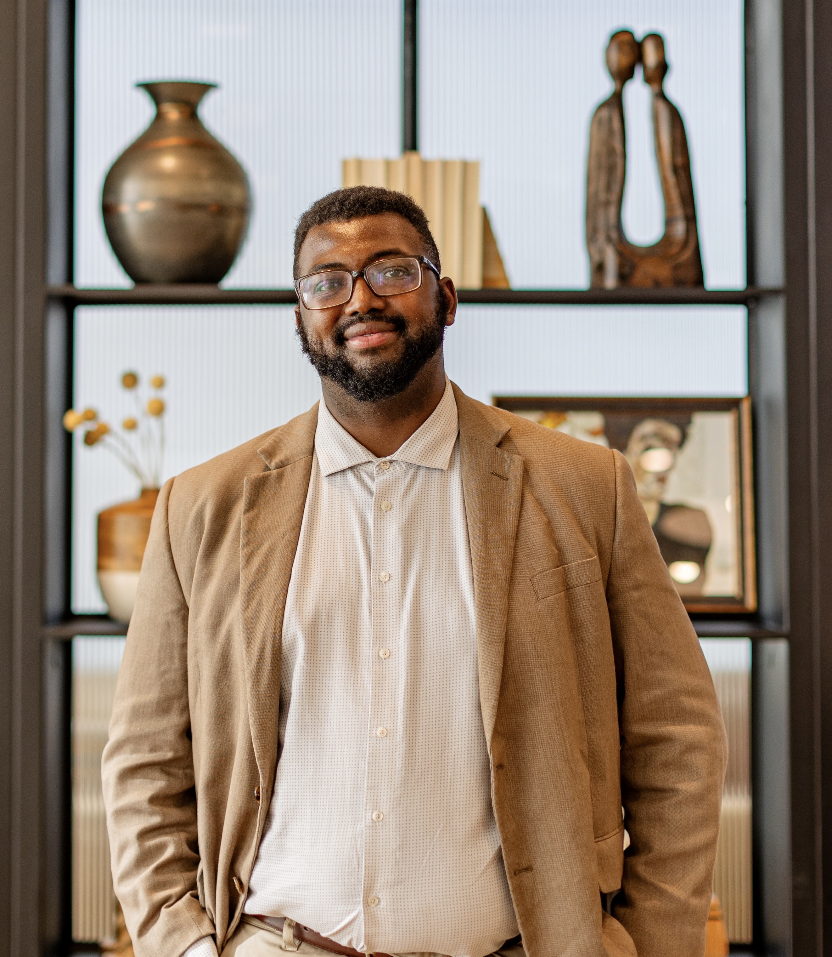 A man in a tan suit stands in front of a shelf with decorative items.