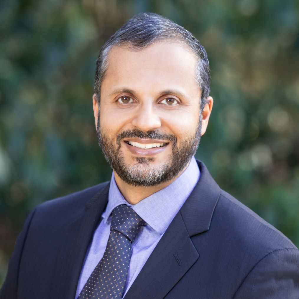 A smiling man in a suit stands outdoors with greenery in the background.