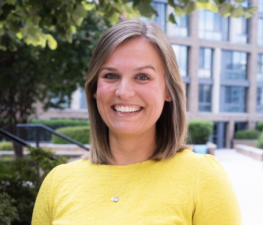 Jenna Miller smiles in a yellow top, outdoors near modern buildings and greenery.