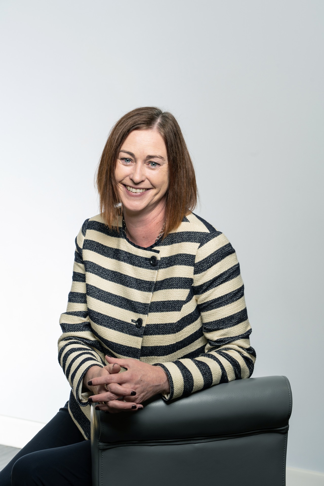 Jo Gavigan smiles while sitting on a chair, wearing a striped blouse against a light backdrop.